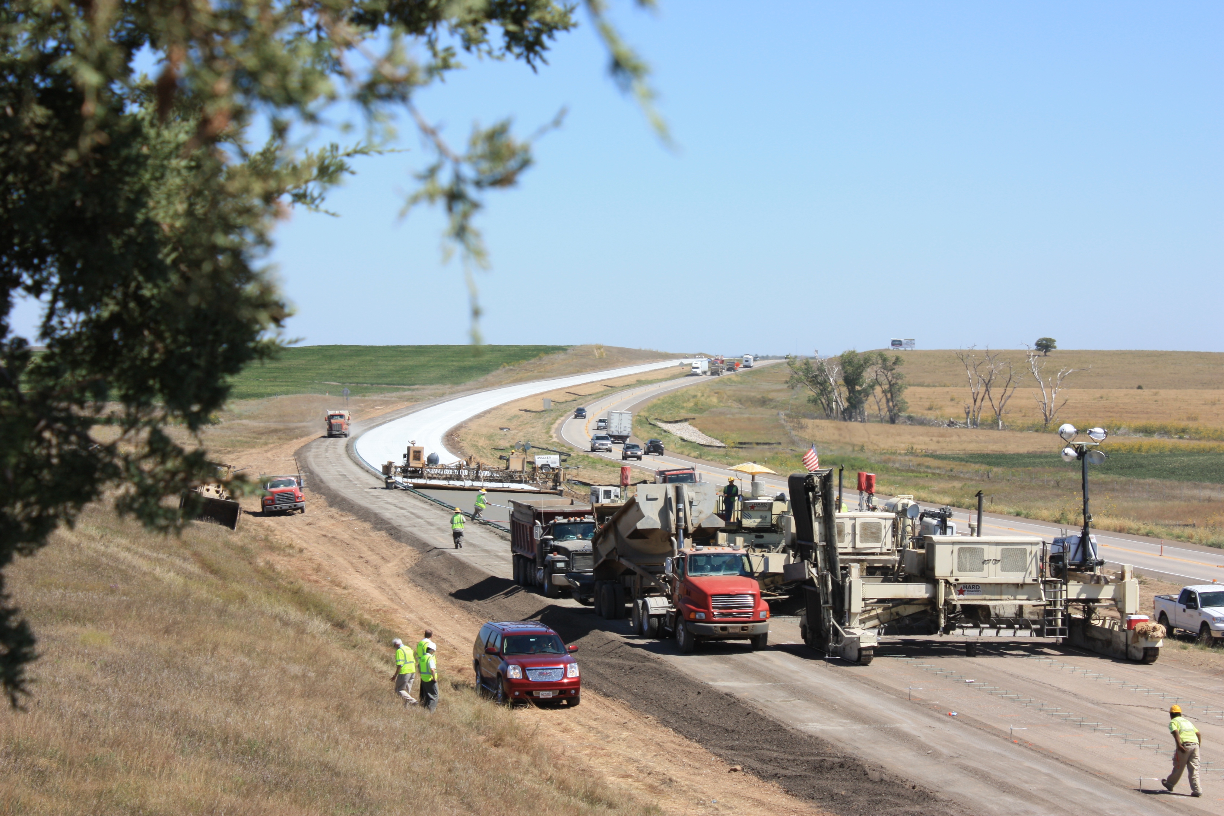 Four-lane divided roadway with lanes at left surrounded by equipment and workers, which are installing a concrete overlay.